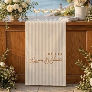 Puede incluir: Un camino de mesa a rayas beige y blancas con el texto impreso TOAST TO Emma & James en una fuente cursiva marr&oacute;n. El camino est&aacute; sobre una barra de madera, acompa&ntilde;ado de cuatro copas transparentes y un arreglo floral blanco.