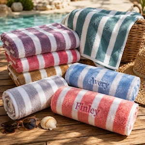 May include: A collection of rolled striped beach towels in various colors including purple, pink, tan, gray, blue, and coral. Two towels feature embroidered names, Avery on a blue towel and Finley on a coral towel, with a seashell and sunglasses.