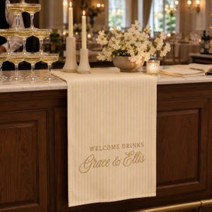 Puede incluir: Un camino de mesa a rayas color crema y blanco con el texto impreso WELCOME DRINKS y los nombres Grace & Ellis en letra cursiva. El camino est&aacute; colocado sobre una barra de madera oscura, bajo dos velas altas y un arreglo floral.