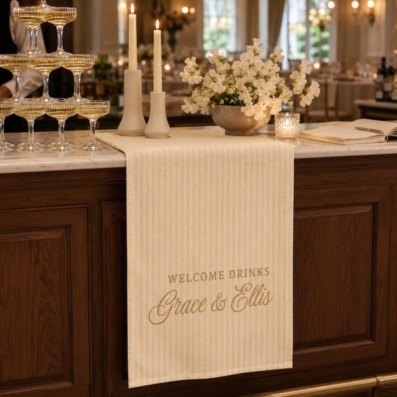 Puede incluir: Un camino de mesa a rayas color crema y blanco con el texto impreso WELCOME DRINKS y los nombres Grace & Ellis en letra cursiva. El camino est&aacute; colocado sobre una barra de madera oscura, bajo dos velas altas y un arreglo floral.