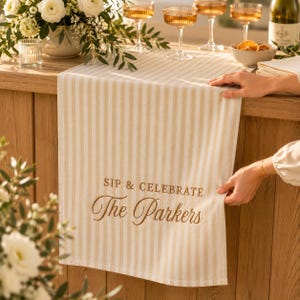 Puede incluir: Un camino de mesa a rayas beige y blancas con el texto bordado SIP & CELEBRATE The Parkers en una fuente marr&oacute;n que combina cursiva y bloque. El camino est&aacute; sobre una superficie de madera, mostrando un patr&oacute;n de rayas verticales.