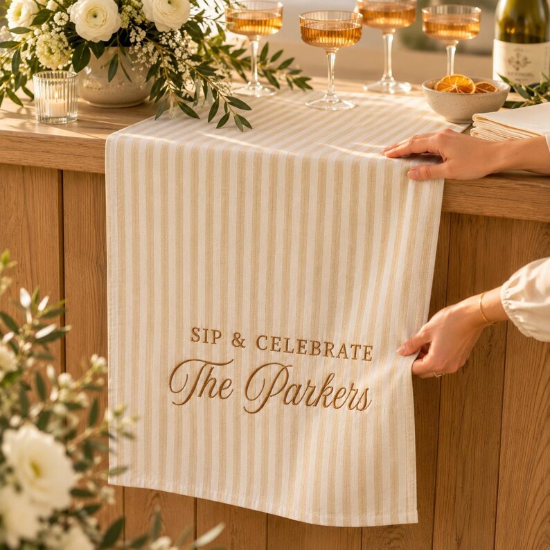 Puede incluir: Un camino de mesa a rayas beige y blancas con el texto bordado SIP & CELEBRATE The Parkers en una fuente marr&oacute;n que combina cursiva y bloque. El camino est&aacute; sobre una superficie de madera, mostrando un patr&oacute;n de rayas verticales.