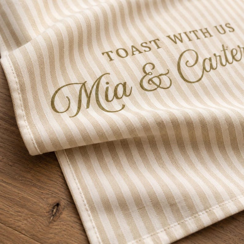 Puede incluir: Un camino de mesa de tela a rayas beige y blancas con el texto impreso TOAST WITH US y los nombres Mia & Carter en una fuente cursiva de color marr&oacute;n oscuro. El tejido tiene una textura visible con bordes cosidos limpios.