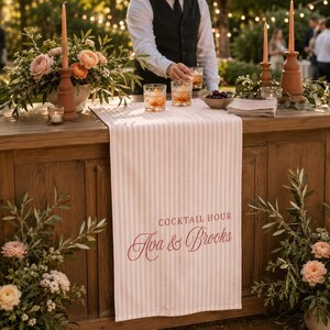 Puede incluir: Un camino de mesa a rayas de color rosa claro y blanco con el texto impreso COCKTAIL HOUR y los nombres Ava & Brooks en una fuente cursiva de color rosa oscuro. El camino est&aacute; sobre una barra de madera decorada con flores y cristaler&iacute;a.