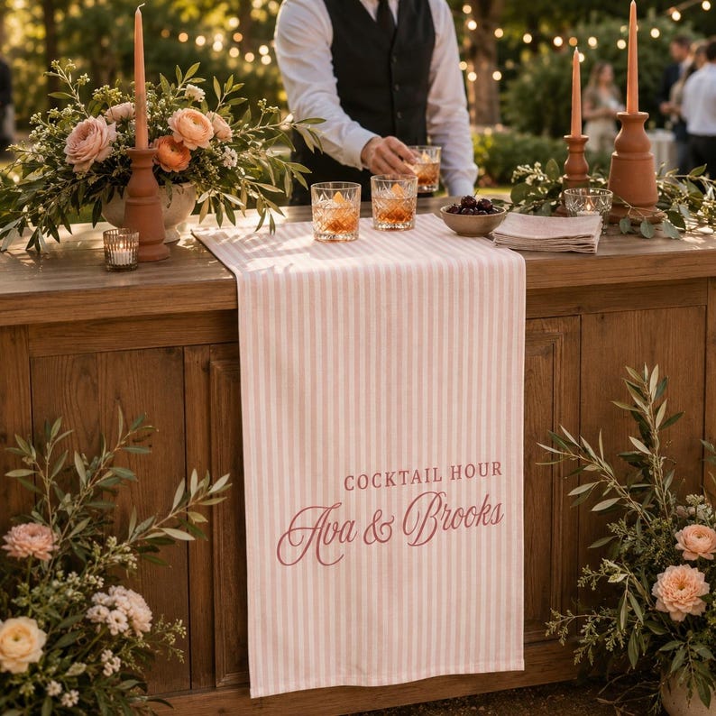 Puede incluir: Un camino de mesa a rayas de color rosa claro y blanco con el texto impreso COCKTAIL HOUR y los nombres Ava & Brooks en una fuente cursiva de color rosa oscuro. El camino est&aacute; sobre una barra de madera decorada con flores y cristaler&iacute;a.