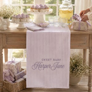 Puede incluir: Un camino de mesa a rayas de color morado claro y blanco con el texto bordado Sweet Baby Harper June en letra cursiva. El camino est&aacute; sobre una mesa de madera con cupcakes en un pedestal blanco, vasos y cajas de regalo.