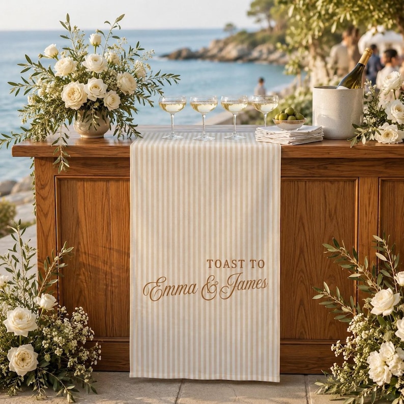 Puede incluir: Un camino de mesa a rayas beige y blancas con el texto impreso TOAST TO Emma & James en una fuente cursiva marr&oacute;n. El camino est&aacute; sobre una barra de madera, acompa&ntilde;ado de cuatro copas transparentes y un arreglo floral blanco.