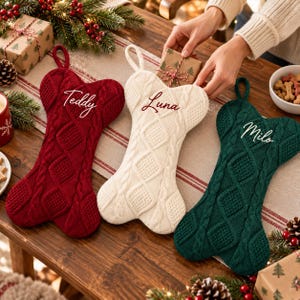 May include: Three bone-shaped Christmas stockings in red, cream, and dark green, each featuring a cable knit pattern and embroidered names. The red stocking is labeled Teddy, the cream stocking is labeled Luna, and the dark green stocking is labeled Milo.