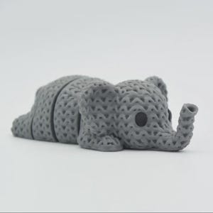 Pode incluir: Estatueta de elefante cinzento texturizado em posição de repouso. O elefante tem orelhas grandes, uma tromba e olhos pretos pequenos. A estatueta é feita de um material com aparência tecida ou de malha, e está sobre uma superfície branca.