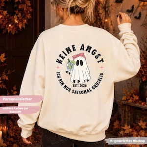 Könnte beinhalten: Ein cremefarbenes Sweatshirt mit Rundhalsausschnitt und einem Rückendruck, der ein Gespenst mit rosa Stirnband zeigt, das einen Cocktail hält. Der Text lautet KEINE ANGST, ICH BIN NUR SAISONAL GRUSELIG und EST. 2026 in kreisförmiger Schrift.