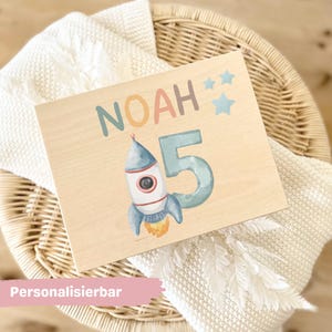 Op de afbeelding: Een rechthoekig houten bord met de naam NOAH in gedempte kleuren, een groot cijfer 5, een illustratie van een raket met een gele vlam en drie lichtblauwe sterren. Het ontwerp is gedrukt op een licht houten oppervlak met een gladde afwerking.