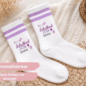 Könnte beinhalten: Ein Paar weiße Socken mit zwei violetten Streifen am oberen Rand. Die Socken sind mit violetten Wimpeln, zwei Luftballons und dem Text Schulkind 2026 Emma in einer Kombination aus Schreibschrift und serifenloser Schrift bedruckt.