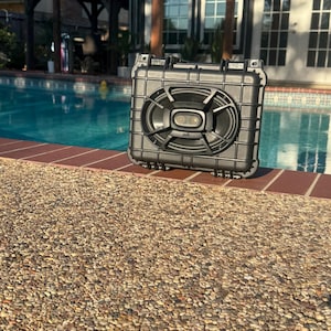 Puede incluir: Un altavoz exterior negro y rectangular con una rejilla de malla y el logotipo de Polk Audio. El altavoz está colocado sobre un borde de ladrillo junto a una piscina. El altavoz es resistente al agua.