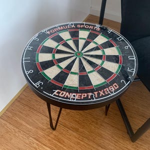 May include: A round side table featuring a dartboard top with black, white, red, and green segments. The text "FORMULA SPORTS" and "CONCEPT X890" is printed on the surface. The table has thin black metal hairpin legs and a circular frame.
