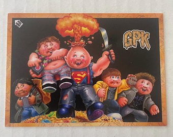 May include: A Garbage Pail Kids trading card featuring several cartoon characters. One character has a head exploding with a mushroom cloud. Other characters are holding a sword, a game controller, and a rock. The card has the letters "GPK" in the upper right corner.