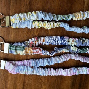 May include: Three fabric lanyards with gathered, scrunchie-style material in various floral and patterned prints. Each lanyard features a silver-toned metal clasp and a circular key ring attached to a rectangular metal connector at the end.