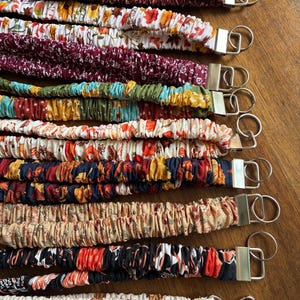 May include: A collection of ten fabric lanyards featuring various patterns including floral, autumn leaves, and geometric prints. Each lanyard is gathered with elastic and finished with a silver-toned metal clasp and a circular key ring attachment.