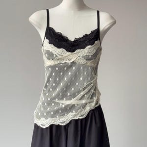 May include: A sleeveless camisole featuring a sheer white mesh fabric with a white polka dot pattern. The garment has a black lace trim along the V-neckline and a white lace trim along the hem, paired with thin black shoulder straps.