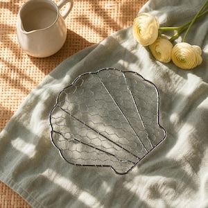 May include: A silver-toned metal wire trivet shaped like a seashell, featuring a fine hexagonal mesh pattern framed by a sturdy wire border. The object has a flat, open design with a scalloped edge, constructed from thin, interconnected metal wires.