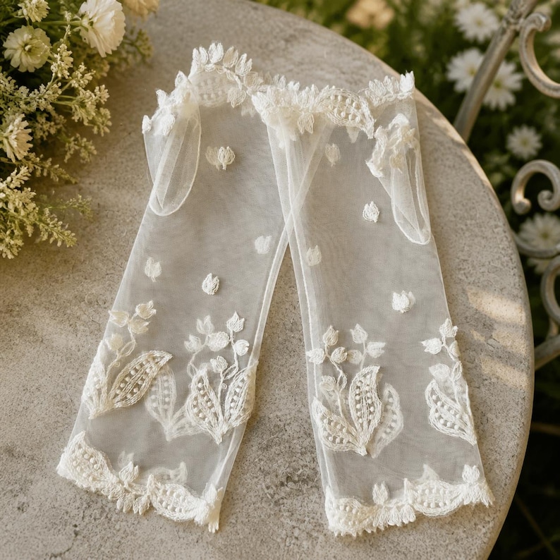 May include: A pair of sheer white fingerless gloves made of fine tulle fabric, featuring delicate white floral embroidery and scalloped lace trim along the top and bottom edges. The gloves are adorned with small, scattered white floral motifs.