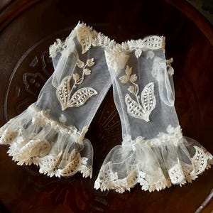 May include: A pair of sheer white fingerless gloves made of fine mesh fabric. Each glove features an embroidered floral motif with delicate leaves and stems, finished with a ruffled, scalloped lace trim at the wrist and the forearm opening.
