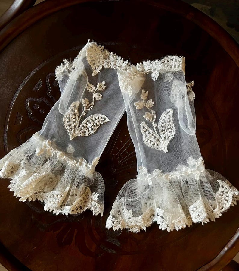 May include: A pair of sheer white fingerless gloves made of fine mesh fabric. Each glove features an embroidered floral motif with delicate leaves and stems, finished with a ruffled, scalloped lace trim at the wrist and the forearm opening.