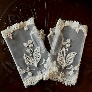 May include: A pair of fingerless gloves made of sheer off-white mesh fabric. Each glove features intricate cream-colored floral embroidery with leaf patterns and a scalloped lace trim along the top and bottom edges, including a thumb hole opening.