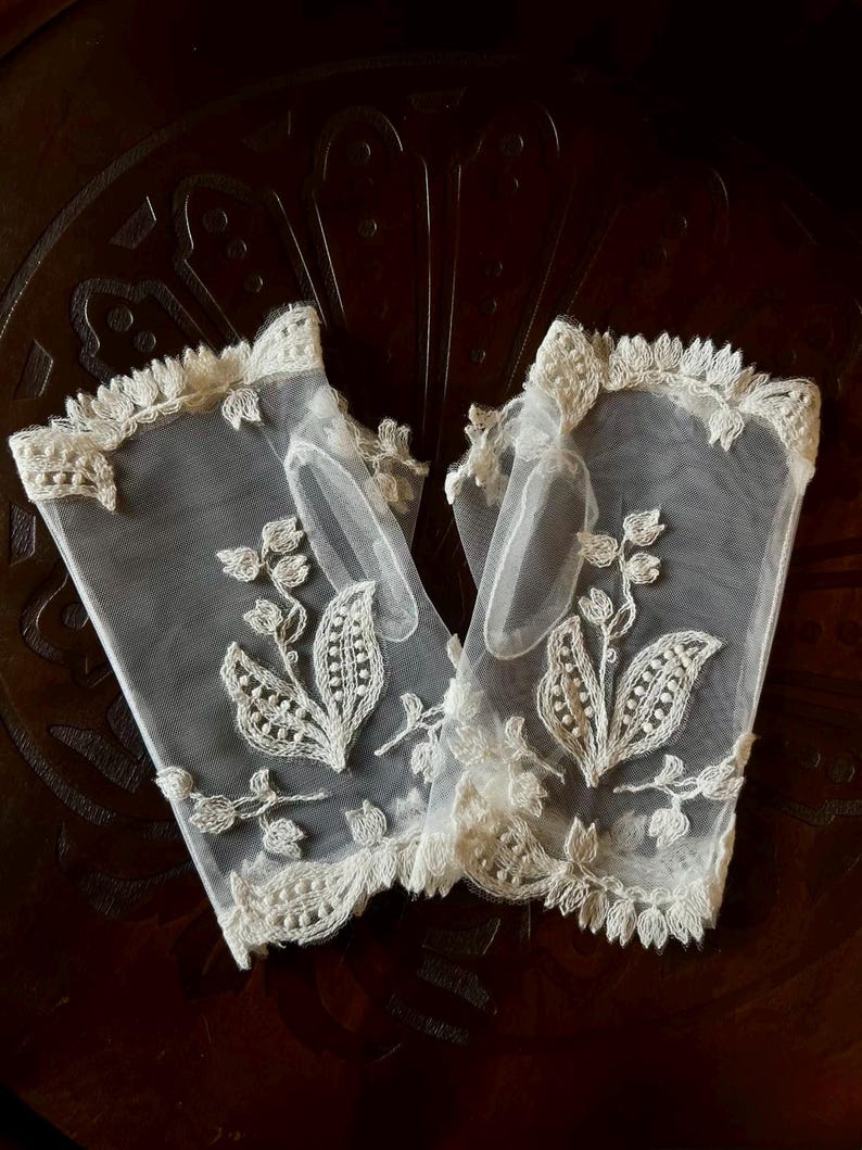 May include: A pair of fingerless gloves made of sheer off-white mesh fabric. Each glove features intricate cream-colored floral embroidery with leaf patterns and a scalloped lace trim along the top and bottom edges, including a thumb hole opening.