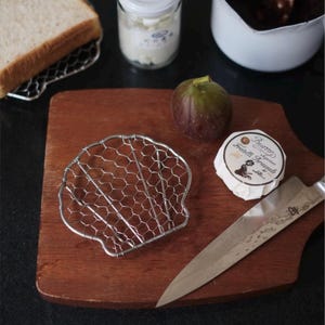 May include: A silver-toned metal wire trivet shaped like a seashell, featuring a hexagonal mesh pattern. The trivet is placed on a wooden cutting board alongside a fresh purple fig, a round butter package labelled Fratelli Brazzale, and a steel knife.