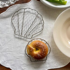 May include: Two silver-toned metal wire racks shaped like seashells. One rack is empty, and the other holds a single round, golden-brown bagel topped with white sesame seeds. The wire racks feature a fine hexagonal mesh pattern within the frame.