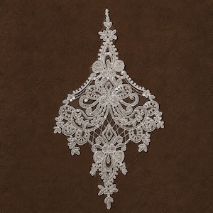May include: A white lace applique featuring an intricate symmetrical design with floral motifs, scrolling filigree patterns, and small clear sequins. The embroidery is worked onto a sheer mesh base, forming a pointed, elongated diamond shape.