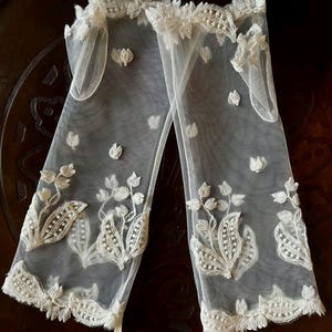 May include: A pair of sheer white tulle gloves featuring intricate ivory floral embroidery and scalloped lace trim along the top and bottom edges. The delicate fabric is adorned with scattered embroidered flower motifs throughout the length.