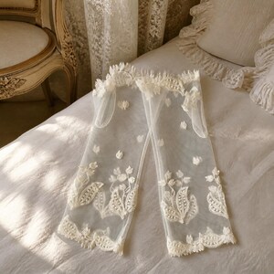 May include: A pair of sheer off-white tulle gloves featuring delicate floral embroidery and scalloped edges. The gloves are crafted from a lightweight mesh fabric with intricate threadwork patterns of leaves and small flowers throughout the length.