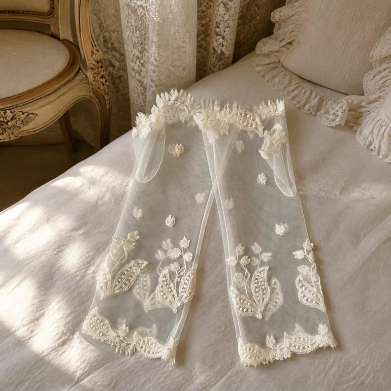 May include: A pair of sheer off-white tulle gloves featuring delicate floral embroidery and scalloped edges. The gloves are crafted from a lightweight mesh fabric with intricate threadwork patterns of leaves and small flowers throughout the length.