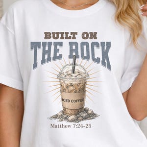 Puede incluir: Camiseta blanca con el texto "BUILT ON THE ROCK" sobre un gráfico de café helado. El gráfico incluye las palabras "ICED COFFEE" y el versículo bíblico Mateo 7:24-25. La camiseta se combina con unos vaqueros azules.