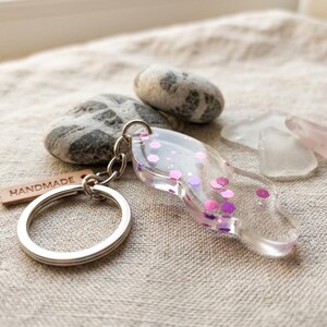 Puede incluir: Un llavero de resina transparente con purpurina rosa y morada. Está unido a un llavero plateado y a una pequeña etiqueta de madera con la inscripción "HANDMADE". El fondo incluye piedras lisas y trozos de vidrio marino.