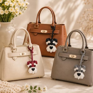 May include: Three structured handbags in cream, tan, and gray, each featuring dual top handles, a gold-tone turn-lock closure, and a hanging charm shaped like a Schnauzer dog with a heart-shaped nose, attached by a matching leather strap.
