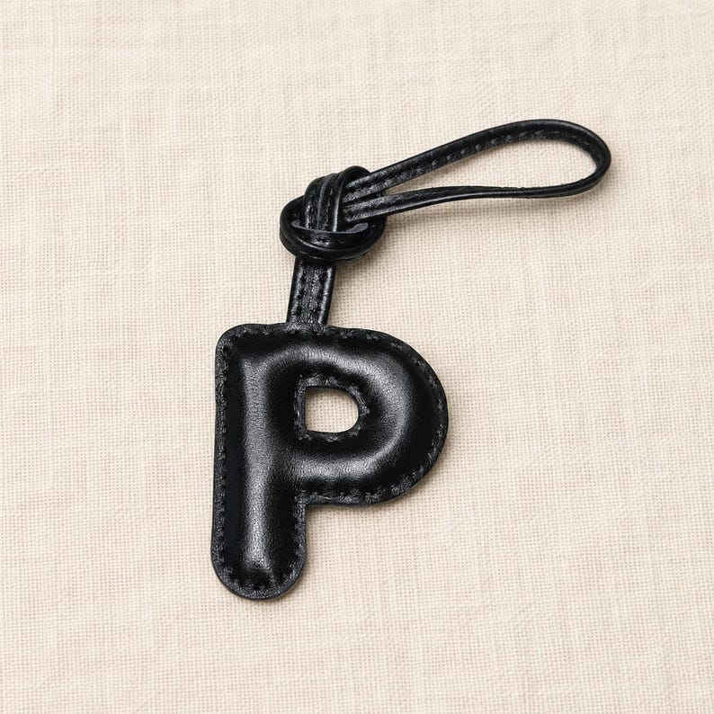 May include: Black leather keychain in the shape of the letter "P". The keychain has a loop for attachment and a stitched border. The letter is a bold, rounded font.