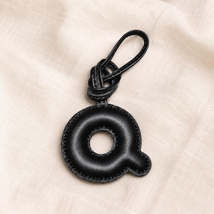 May include: A black leather keychain in the shape of the letter "Q". The letter is stitched around the edges and has a loop at the top for attaching to keys. The keychain is made of a smooth, dark material.
