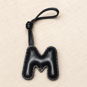 May include: Black leather keychain in the shape of the letter "M". The letter is stitched around the edges with a lighter thread. The keychain has a loop at the top for attaching to keys or a bag.