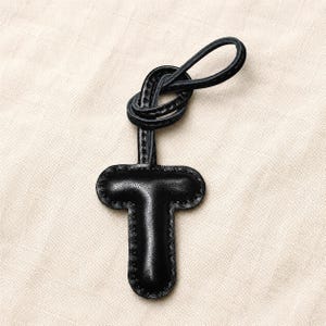 May include: A black leather key fob in the shape of the letter "T". The fob has a loop at the top and is stitched around the edges. The background is a light beige fabric.