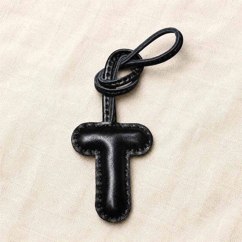 May include: A black leather key fob in the shape of the letter "T". The fob has a loop at the top and is stitched around the edges. The background is a light beige fabric.