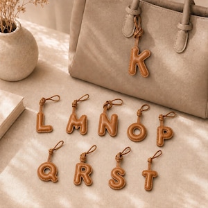 May include: A collection of tan leather-like alphabet letter charms, each featuring a puffy, stitched design and a small loop for attachment. One charm is attached to a tan handbag, while the letters L, M, N, O, P, Q, R, S, and T are arranged below.