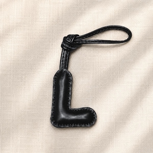 May include: Black leather letter "L" keychain with a knotted loop. The letter is stitched around the edges with a contrasting thread. The keychain is designed to be attached to a bag or set of keys.