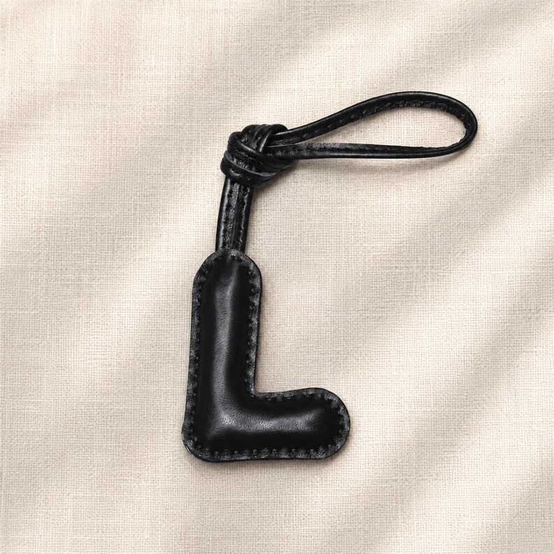May include: Black leather letter "L" keychain with a knotted loop. The letter is stitched around the edges with a contrasting thread. The keychain is designed to be attached to a bag or set of keys.