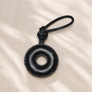 May include: A black leather keychain with a circular design and a loop for attaching keys. The keychain has a donut-like shape with a hole in the center and is stitched around the edges. The loop is also made of black leather.