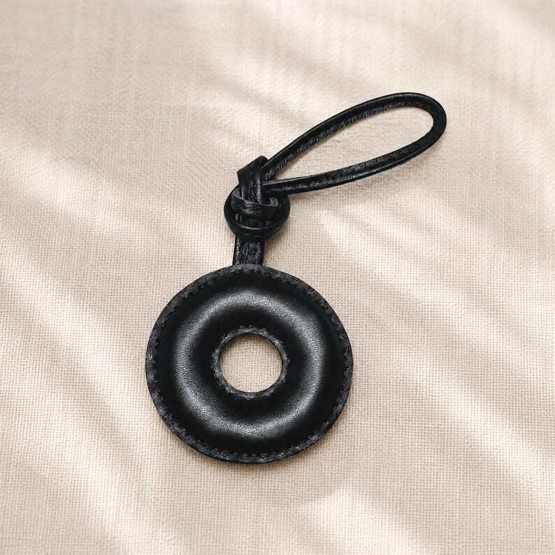 May include: A black leather keychain with a circular design and a loop for attaching keys. The keychain has a donut-like shape with a hole in the center and is stitched around the edges. The loop is also made of black leather.
