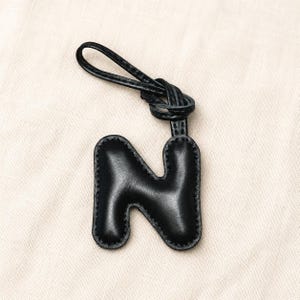May include: Black leather letter "N" keychain with a knotted loop. The letter is stitched around the edges and has a smooth, shiny finish. The keychain is a personalized accessory.