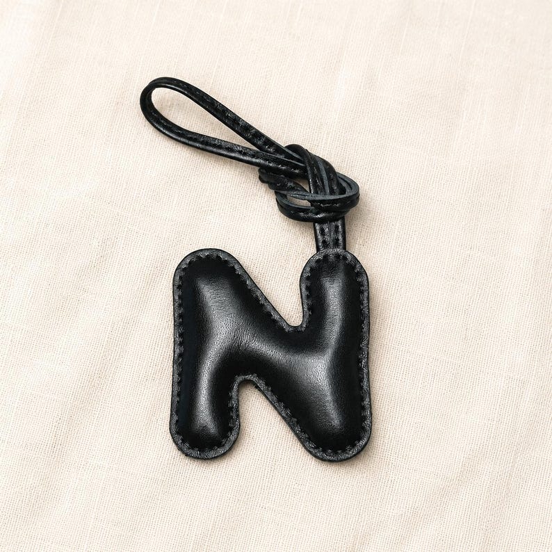 May include: Black leather letter "N" keychain with a knotted loop. The letter is stitched around the edges and has a smooth, shiny finish. The keychain is a personalized accessory.