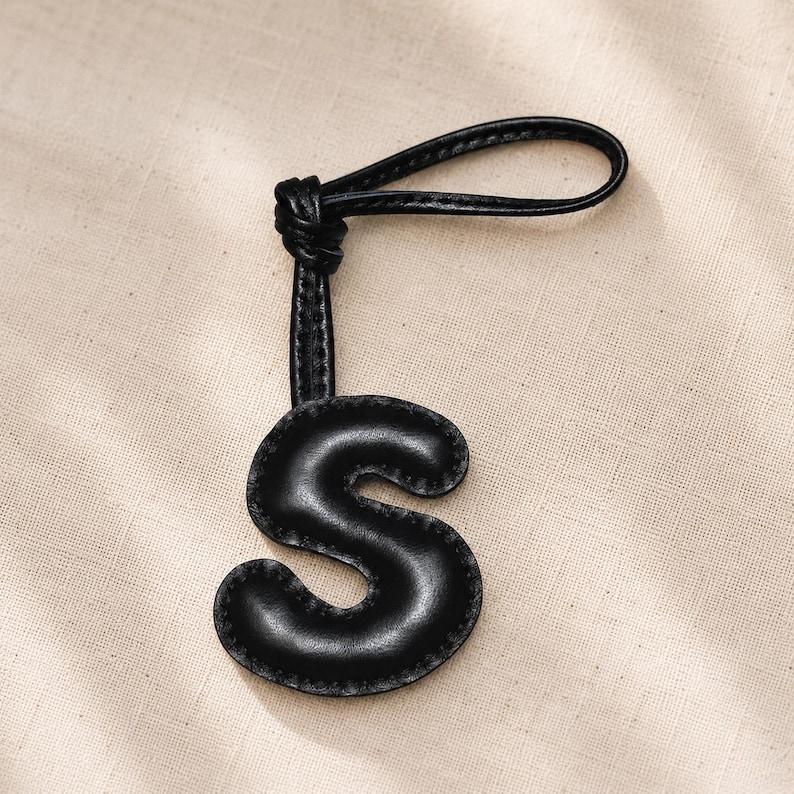 May include: Black leather letter "S" keychain with a loop. The letter is stitched around the edges and attached to a knotted strap. The keychain is set against a neutral background.